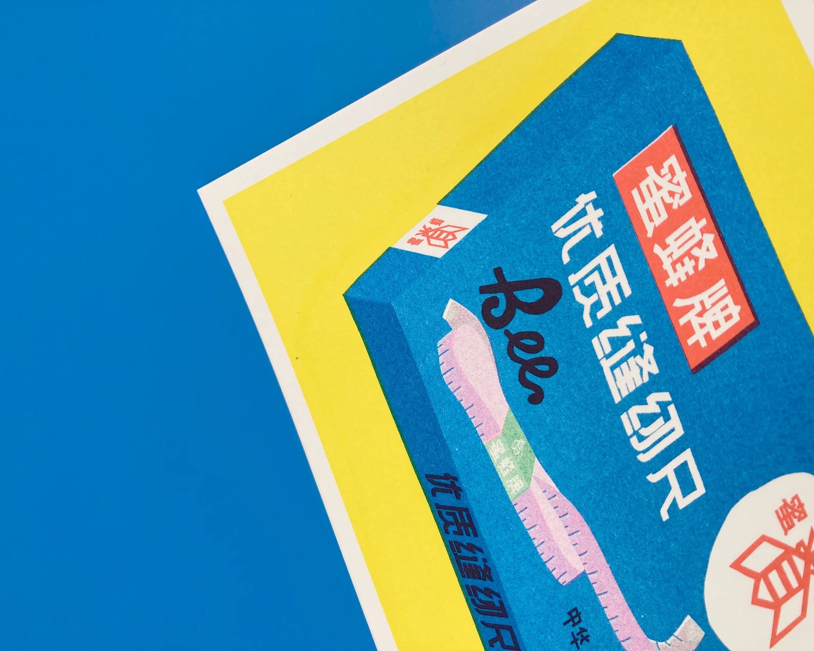 Risograph print of a Chinese Bee brand cloth measuring tape box in bright blue and yellow, created by We Are Out of Office – retro sewing supplies artwork on 300g Biotop paper.