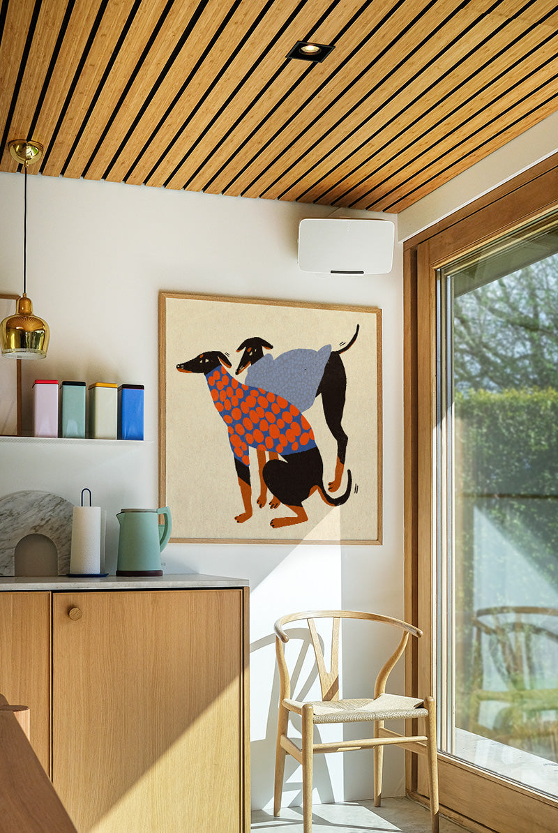 Colorful Doggos art print by Enikő Eged featuring two stylized black dogs wearing patterned sweaters in blue, orange, and grey tones on a warm neutral background, square contemporary giclée wall art for modern interiors.