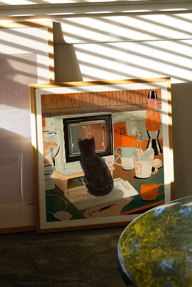 Retro Gaming Cat art print by Enikő Eged, featuring a black cat sitting on a desk in front of a vintage computer game, surrounded by retro home decor, warm orange tones, a lava lamp, coffee cup, and playful nostalgic details.