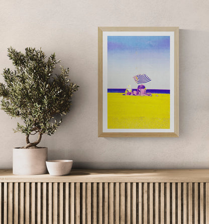 Colorful risograph art print titled “Basque Beach” by PTRVRKD, featuring two people sitting under a striped umbrella on a bright yellow beach with a soft blue-purple sky; A3 limited edition printed on Munken Pure Rough paper. Vibrant modern beach artwork ideal for coastal decor and contemporary wall art collectors.