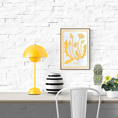 Handmade yellow linocut art print by Belgian artist Leda Vaes titled “Rise,” featuring stylized dandelion flowers and leaves carved in a bold, organic design; A4 nature-inspired print with warm, uplifting botanical energy.