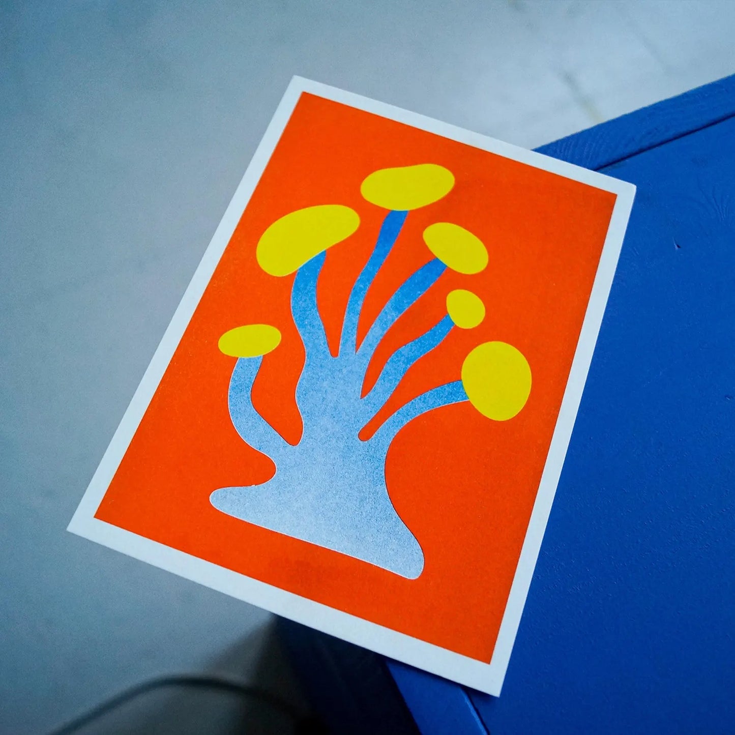 Blue and yellow funky mushroom risograph print by Dutch illustrator PTRVRKD – playful abstract botanical design with organic blue stems, bold yellow caps, and fiery orange background, printed with eco-friendly soy inks on 250 gsm Biotop paper, handmade in the Netherlands – quirky small art print for modern interiors.
