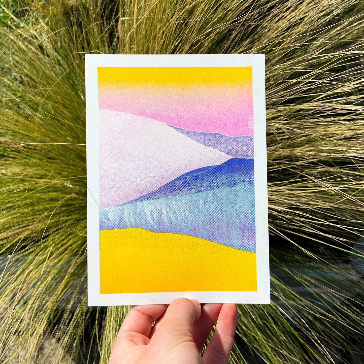 Minimal pastel landscape risograph print by Dutch illustrator PTRVRKD featuring yellow sand dunes, blue hills, and a soft pink sky gradient. Handmade 12.5 x 17.5 cm art print on Biotop 250 gsm paper with soy-based inks. Ideal wall art for modern interiors, Scandinavian style, or desert-inspired home décor.