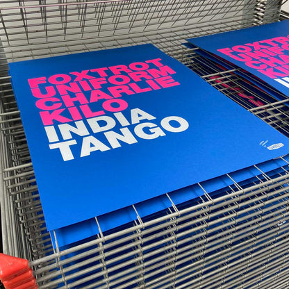 Large blue screen print with bold pink and white typography spelling 'Foxtrot Uniform Charlie Kilo India Tango' by Dutch artist Gumbootz — modern statement art poster, ideal for contemporary spaces.